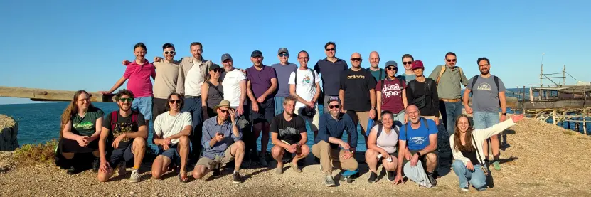 Group photo of Django on the Med 2026 participants during a bike ride on the Adriatic cycle path, along the former Adriatic railway line near the Trabocchi Coast in Abruzzo. The group is posing by the sea in the warm afternoon sunset light, with the Adriatic Sea behind them and the Punta Isola trabocco visible on the right.