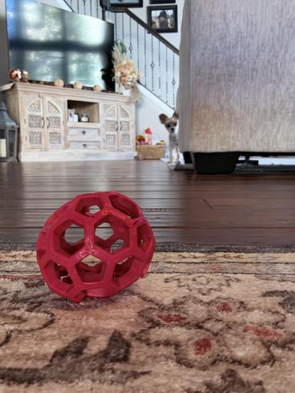 A red hollow ball on a patterned rug; a small dog peeks from behind a sofa near a cabinet and staircase.