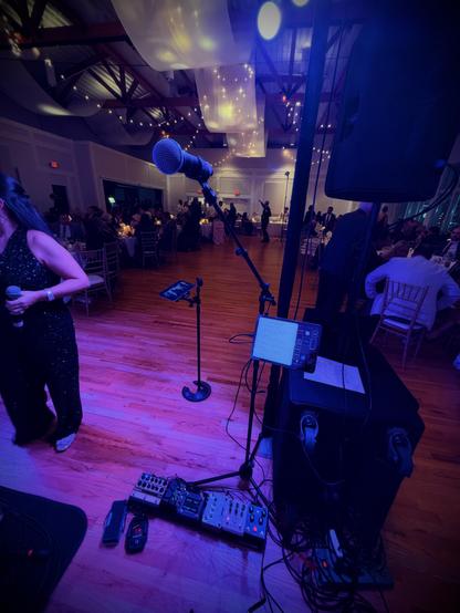 A woman holds a mic near a mic stand with pedals and a tablet, guests sit at tables under glowing lights in a hall.