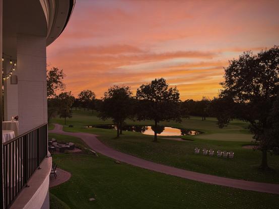 Balcony with string lights overlooks a winding path, pond, trees, and a vivid orange sunset sky across a green golf course.