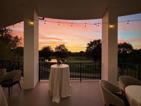 Balcony with string lights, white table with flowers, chairs, overlooking golf course and pond under orange sunset.