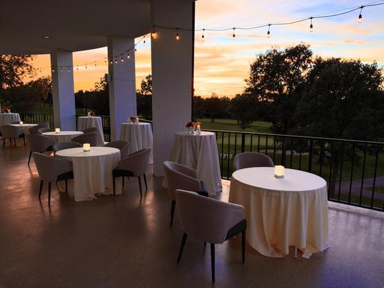 Covered patio with string lights, white tables, chairs, overlooking golf course and trees under orange sunset.