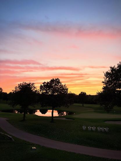 A sunset with pink and orange clouds over a golf course with silhouetted trees, a pond, a path, and six chairs.