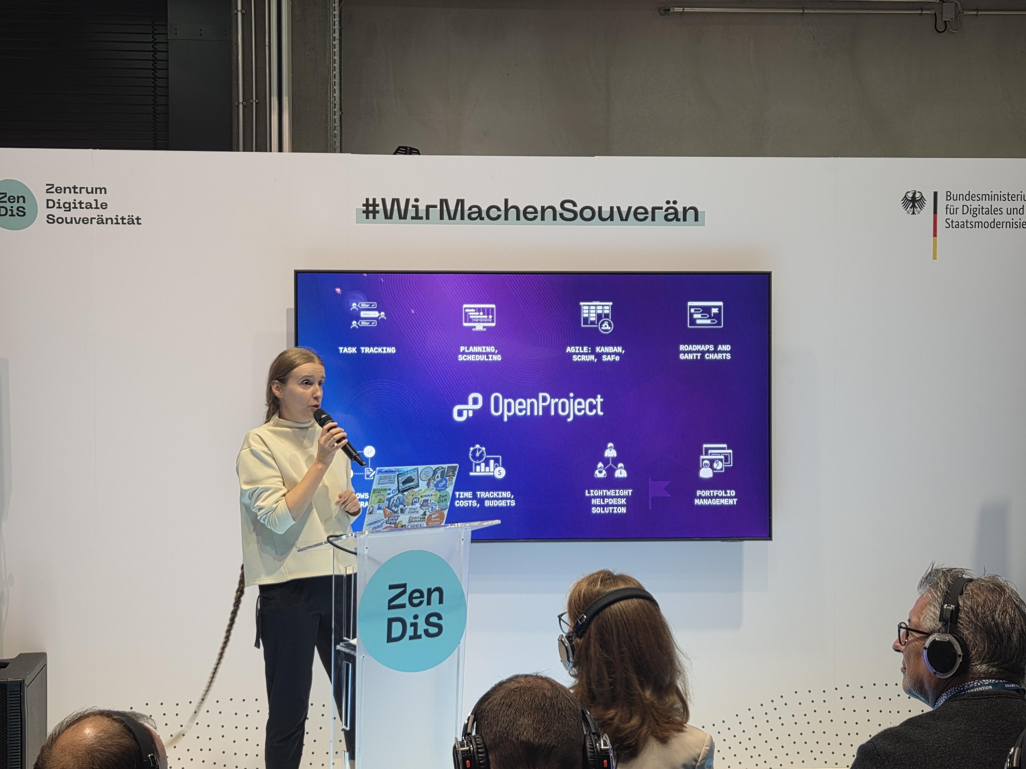 A woman speaks at a podium during a presentation at an event, with a large screen behind her displaying information about project management tools, including task tracking, planning, and scheduling. Attendees wearing headphones are seated in front of her.