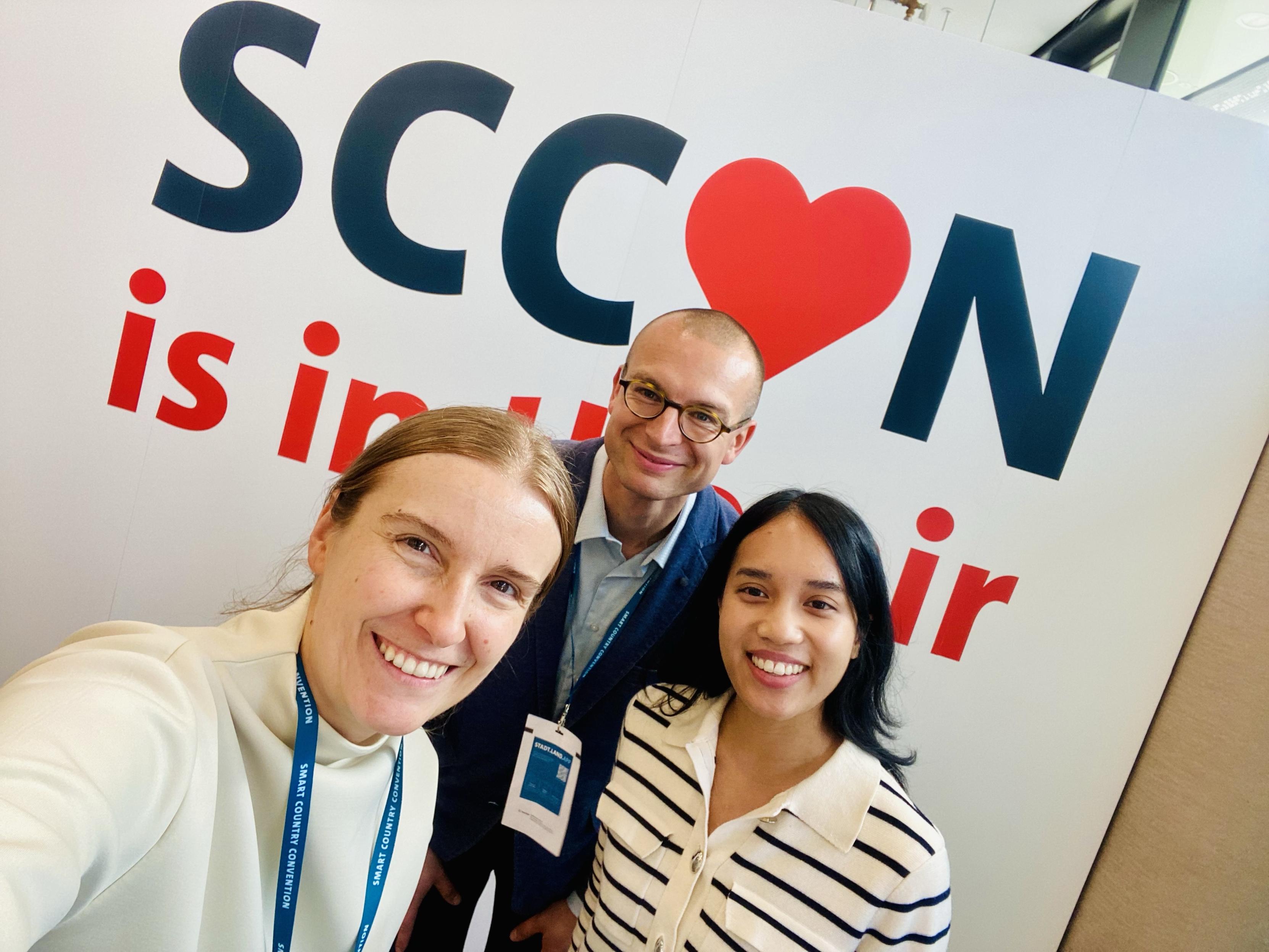Three individuals are posing for a selfie in front of a large sign that reads "SCCON is in the air." The background features prominently displayed text with a heart symbol incorporated into the name.