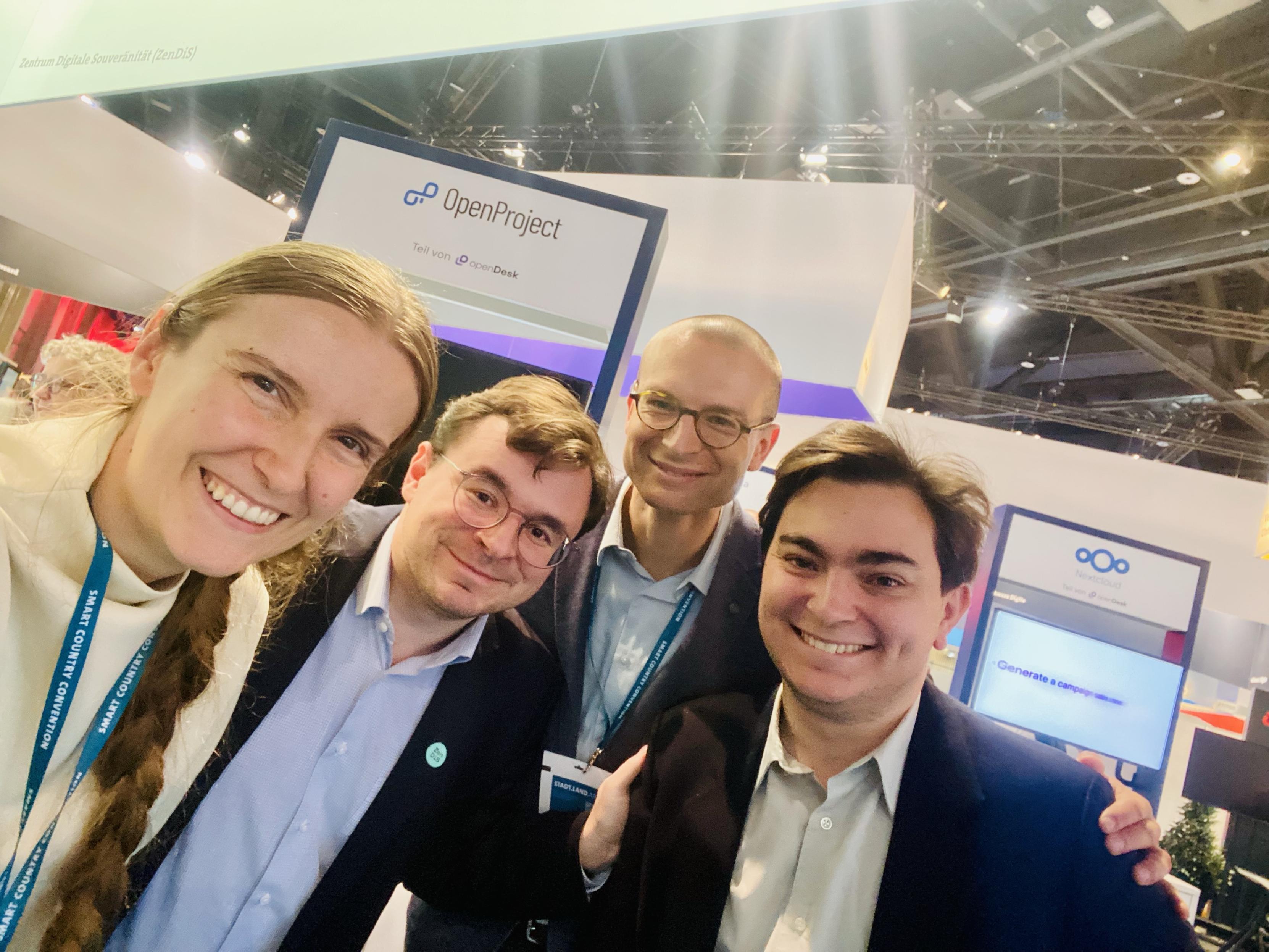 A group of four professionals poses for a selfie at a conference booth. They are smiling and appear engaged, with promotional signage in the background related to "OpenProject."