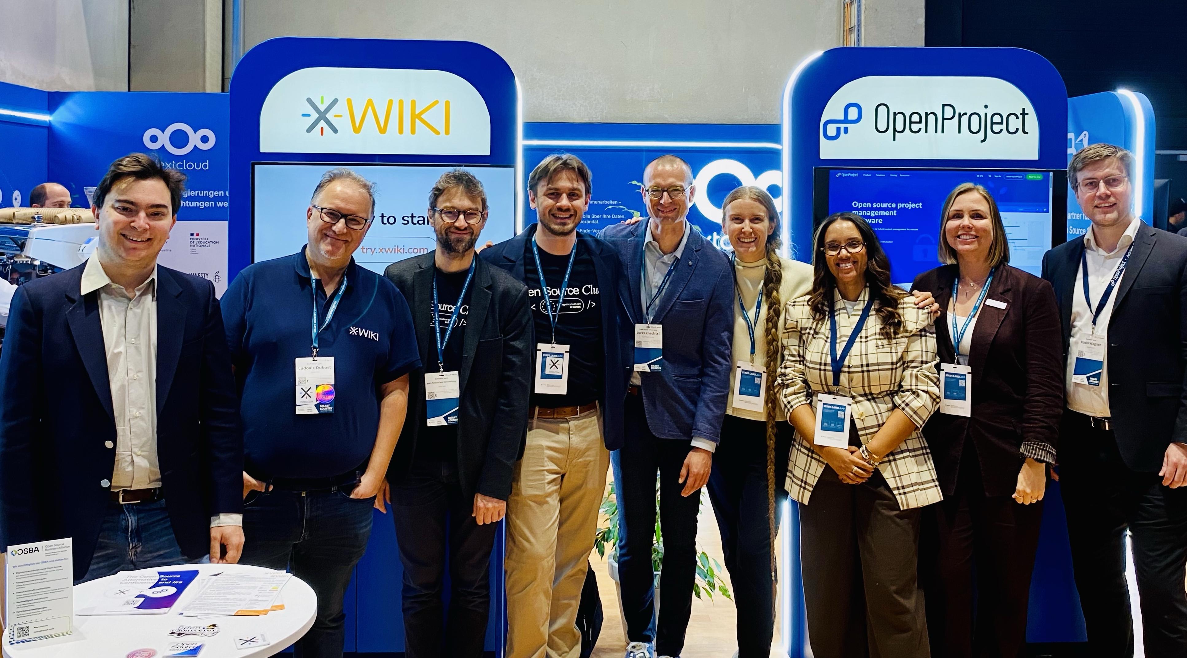 A group of nine people is posing in front of booths featuring the logos for xWiki and OpenProject at a conference or event. The participants are dressed in business attire, some wearing name tags. The background includes promotional materials and a vibrant display.