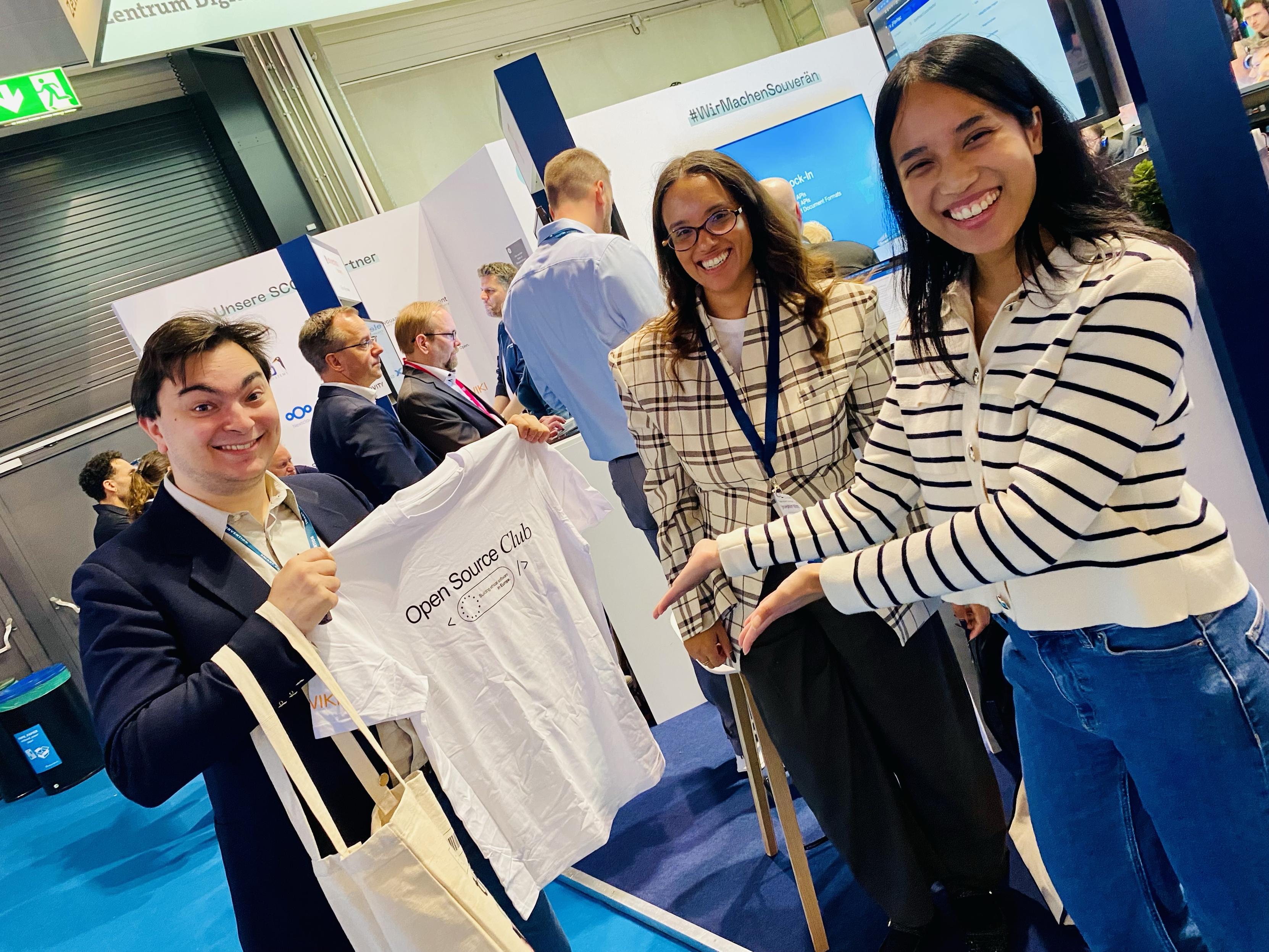 Three people are smiling at a networking event. One person is holding a white T-shirt with "Open Source Club" printed on it. The others are presenting the shirt, and there are other attendees in the background. The setting appears to be a conference