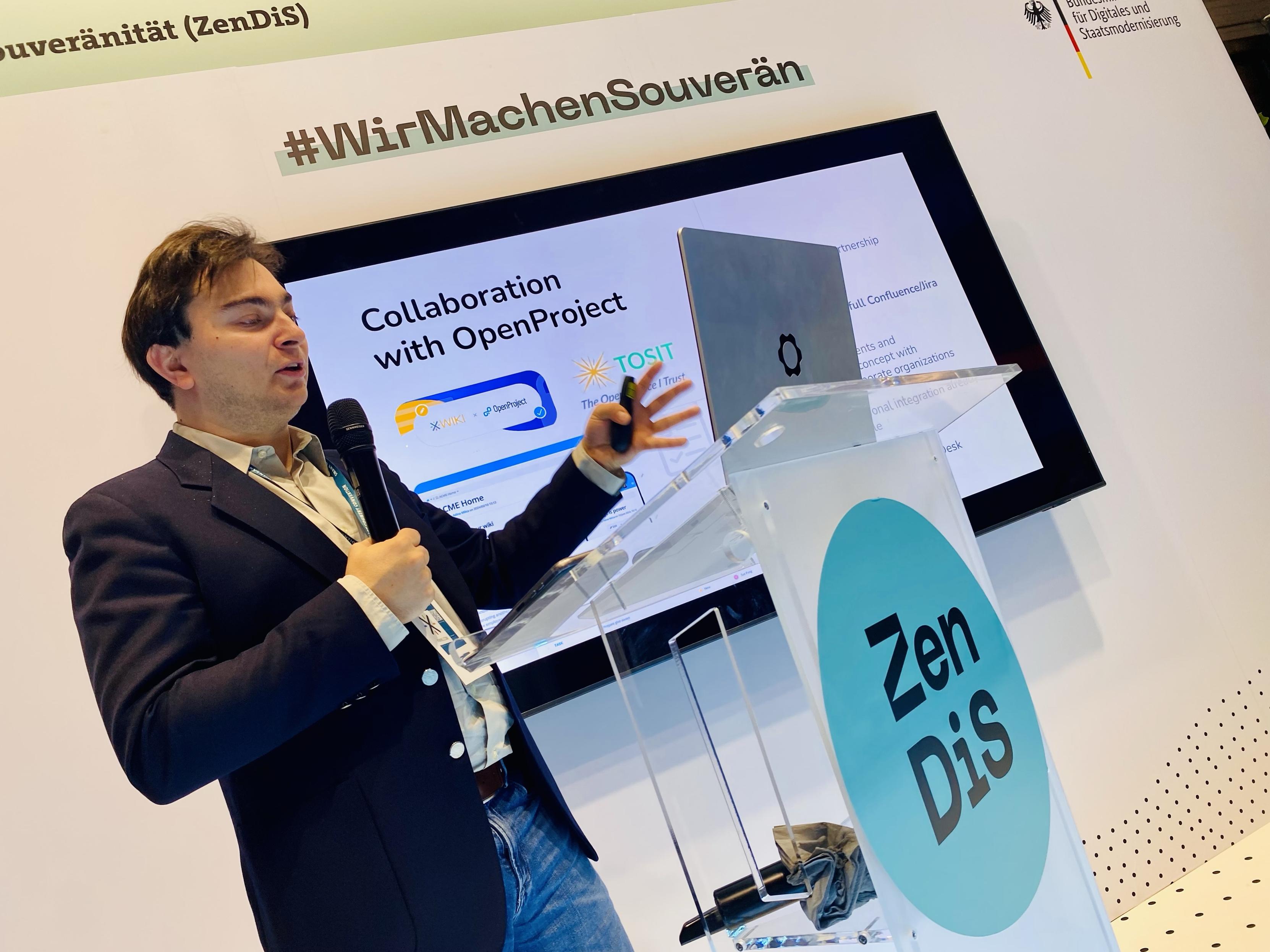 A speaker presents information on collaboration with OpenProject at an event. Behind the speaker is a screen displaying content related to the presentation. The podium features the logo "ZenDis," and there is a hashtag "#WirMachenSouverän" visible.