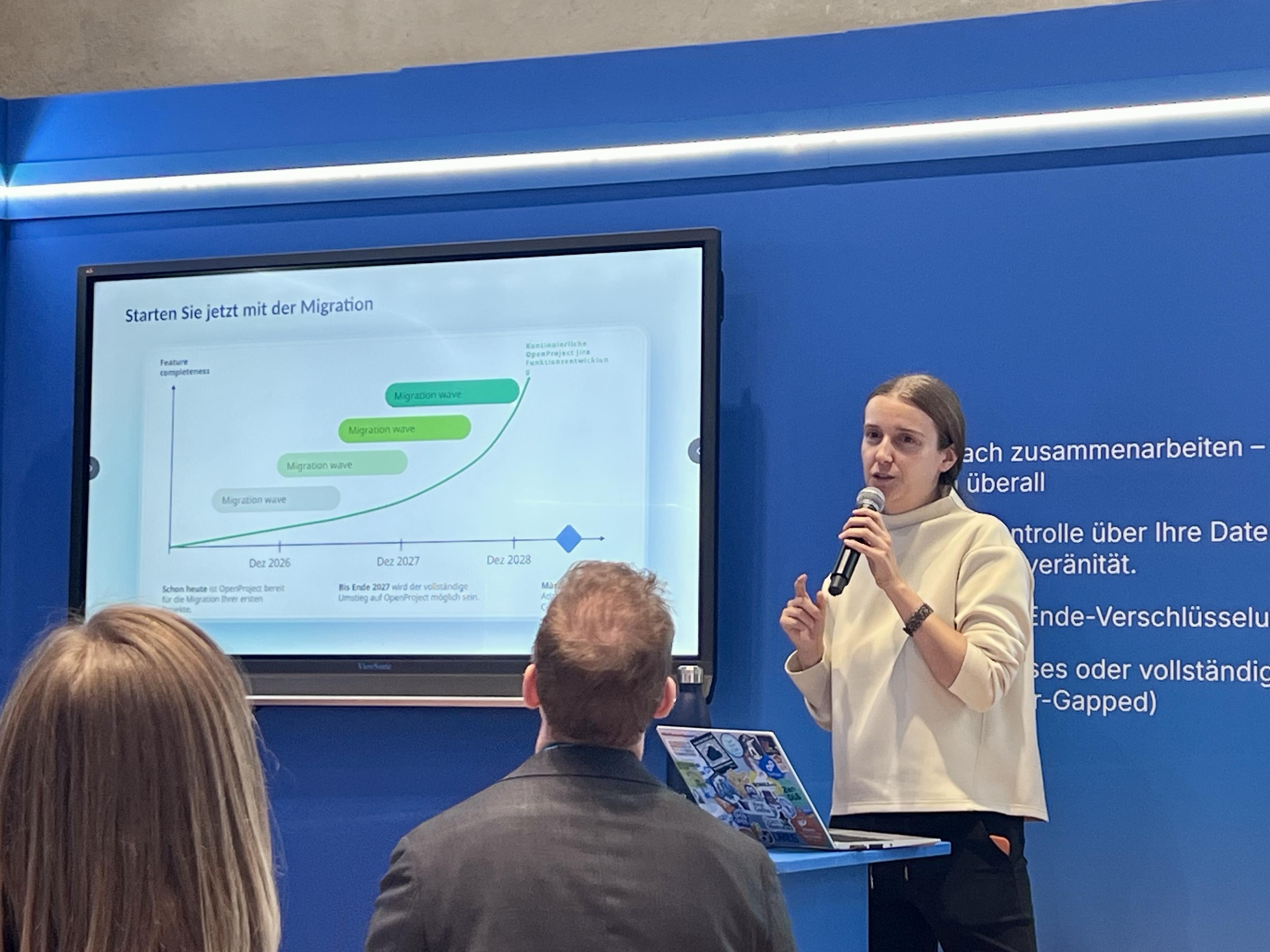 A speaker presents in front of a screen displaying a graph about migration waves with a timeline extending to 2028. The background features a blue wall with text in German. An audience member is visible, focused on the presentation.