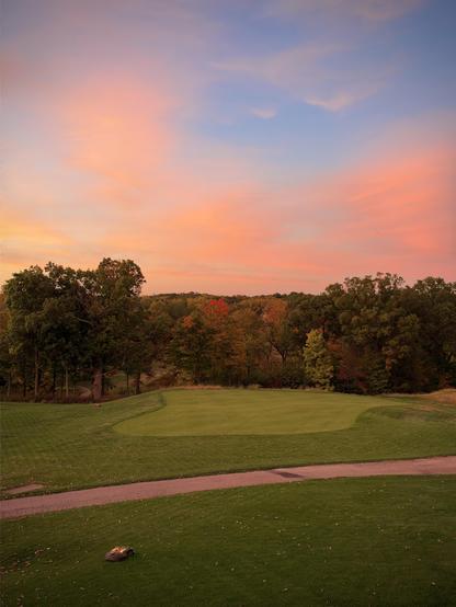 A golf green under a pink and blue sunset sky, bordered by autumn trees and a dirt path.