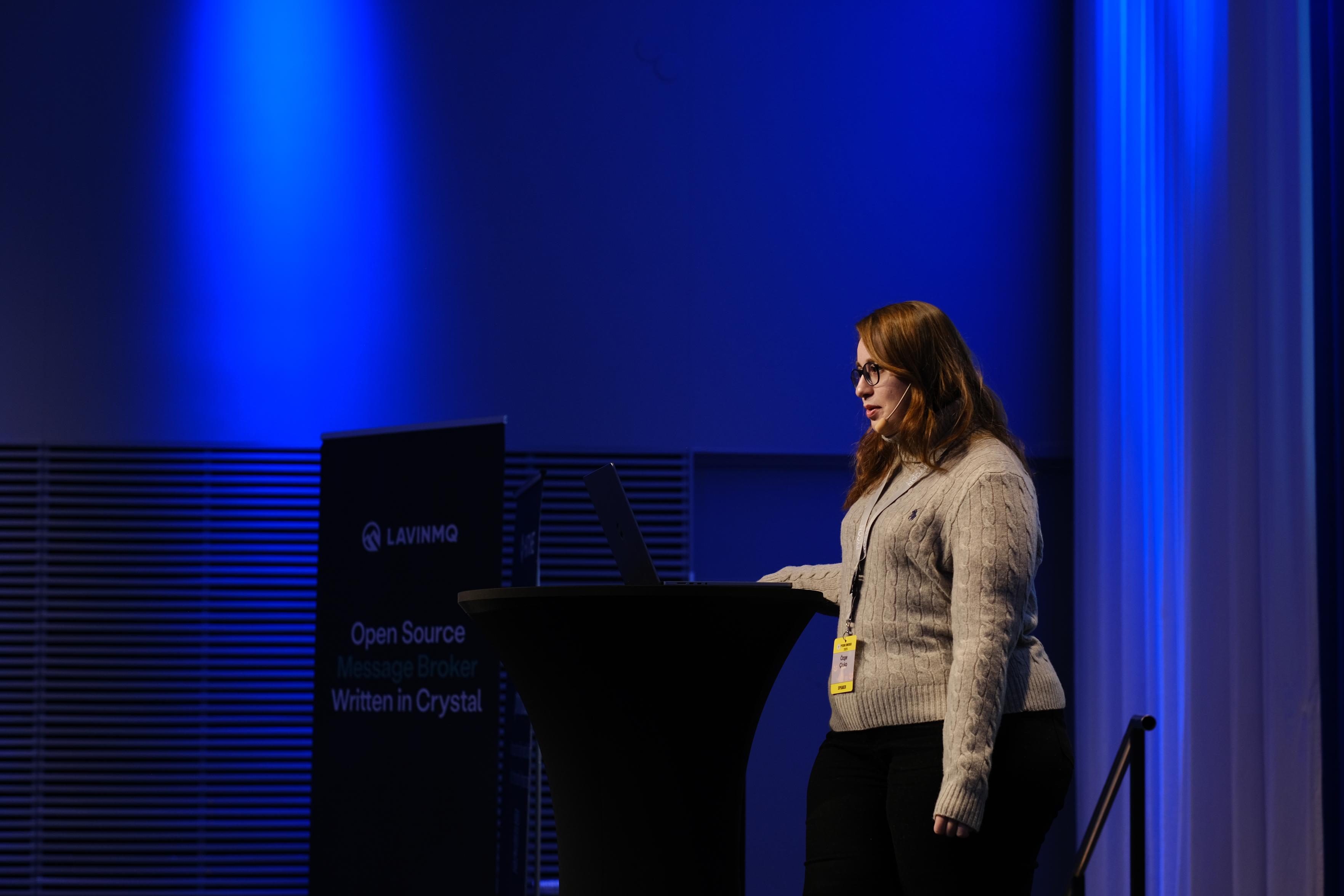 Based on the image provided, here is a description:
**Subject and Action**
A woman with long reddish-brown hair stands at a black podium, appearing to give a presentation or speech. She is looking down at a laptop screen placed on the podium. She is wearing glasses, a light grey cable-knit sweater, dark pants, and a headset microphone. Around her neck is a lanyard with a yellow conference badge.
**Setting and Background**
The setting appears to be a conference stage. The background is dominate