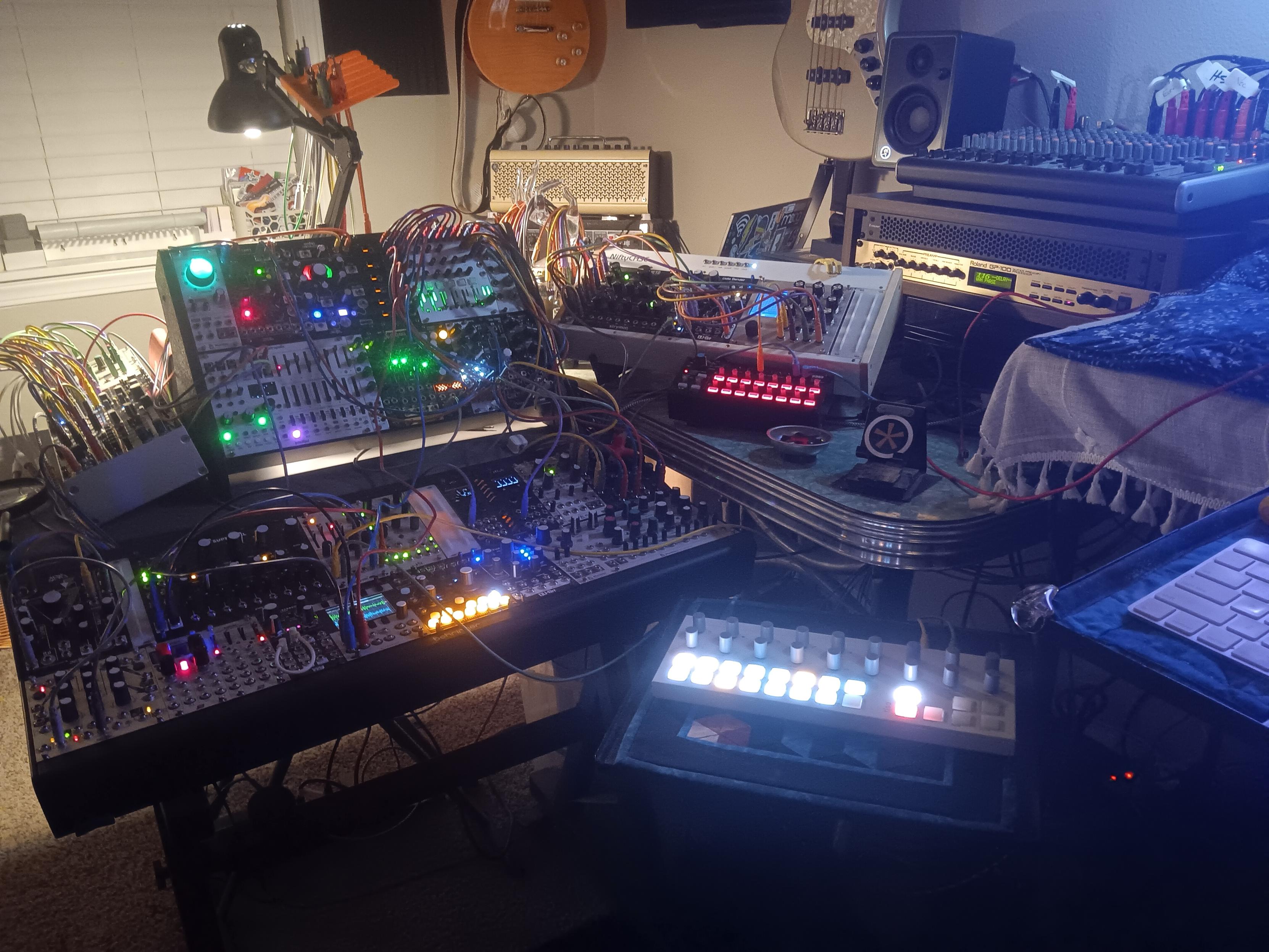 Modular gear of my music studio: 4-part eurorack - modular drums, synths, and lots of effects. A little black (with red knob lights) Korg SQ-1 sequencer. A Torso T-1 algorithmic sequencer (with the bright pad lights). A couple guitars are visible, hanging on the wall. A tan, Yamaha THR30ii amp. A GP-100 effects processor in the rack, with my mixer sitting above.