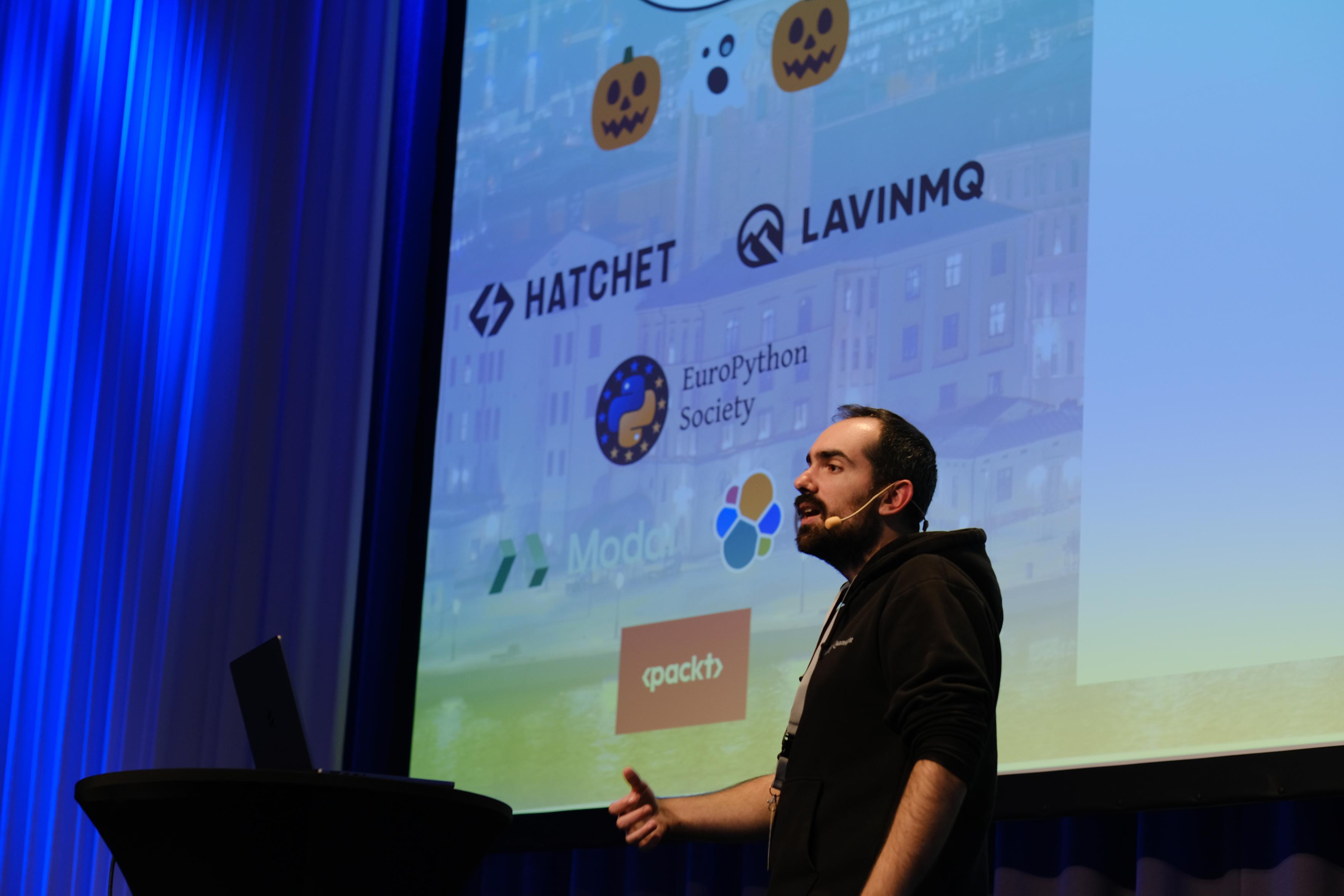 Based on the image, here is a description:
**Subject and Action**
A man with dark hair and a beard is standing on a stage, giving a presentation. He is wearing a black hoodie and a headset microphone. He is facing slightly to the left, gesturing with his right hand as if explaining a point to an audience. In front of him (to the left) is a black podium with a laptop open on it.
**Background and Screen**
Behind the speaker is a large projection screen displaying a slide. The slide features seve