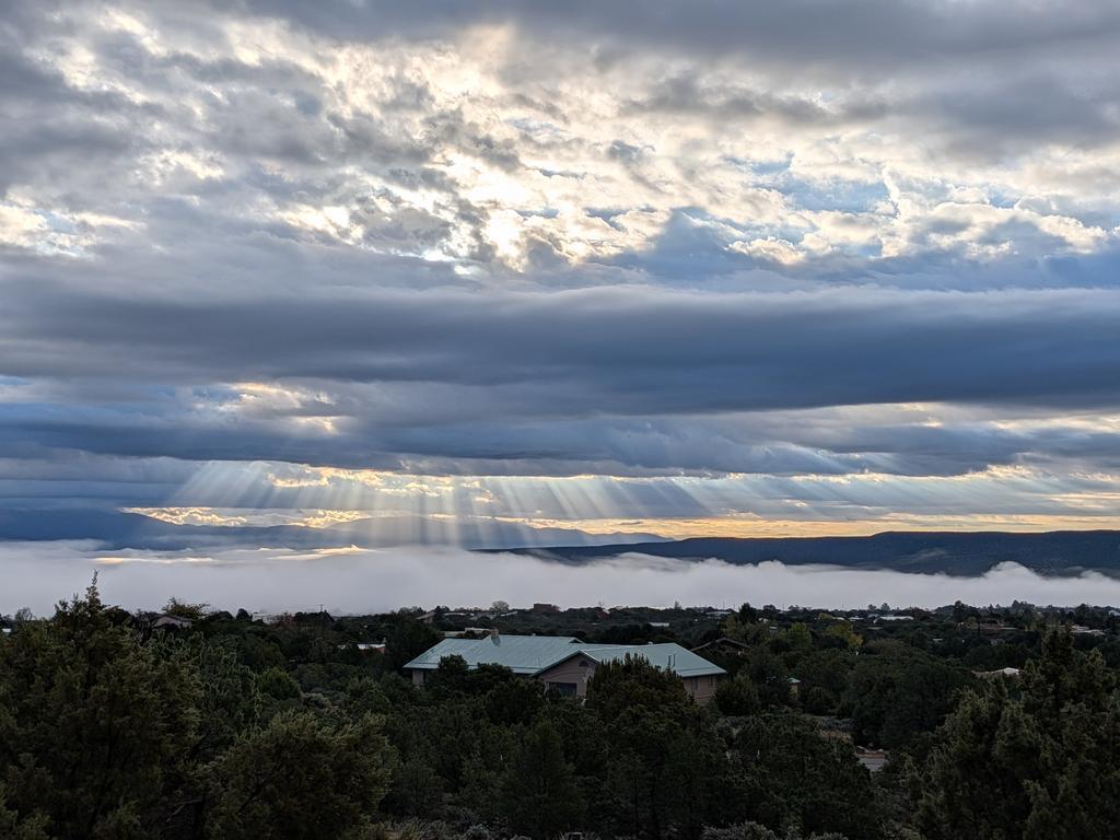 A sunrise sky with lots of cumulus clouds, and crepuscular rays ("god rays") coming through, shining down on the roof of a house that's otherwise obscured by trees.