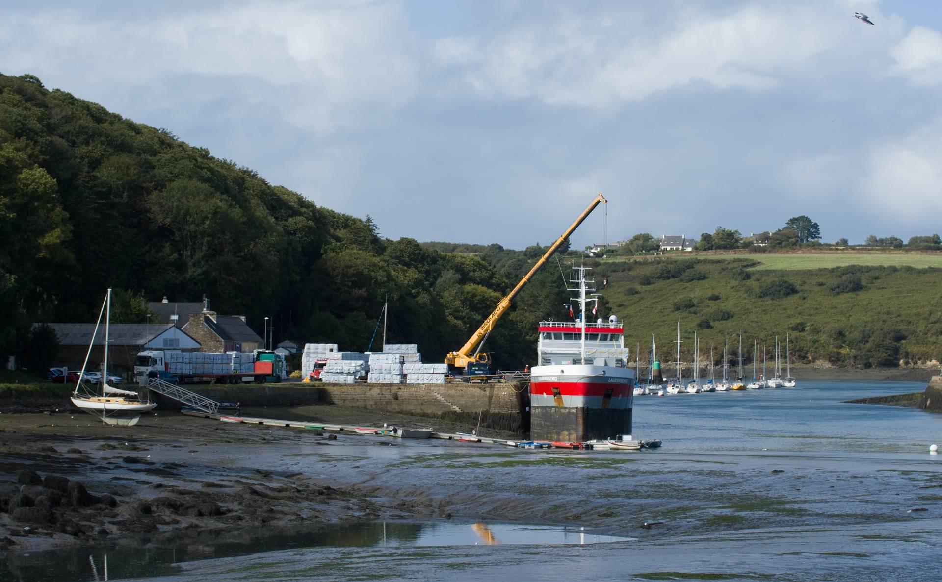 MV Laurierborg seen head-on, high-and-dry. To the left of her the pier as described in the post. In the foreground the river bottom exposed by the low tide. In the background wooded hills rise towards a cloudy and pale blue sky.