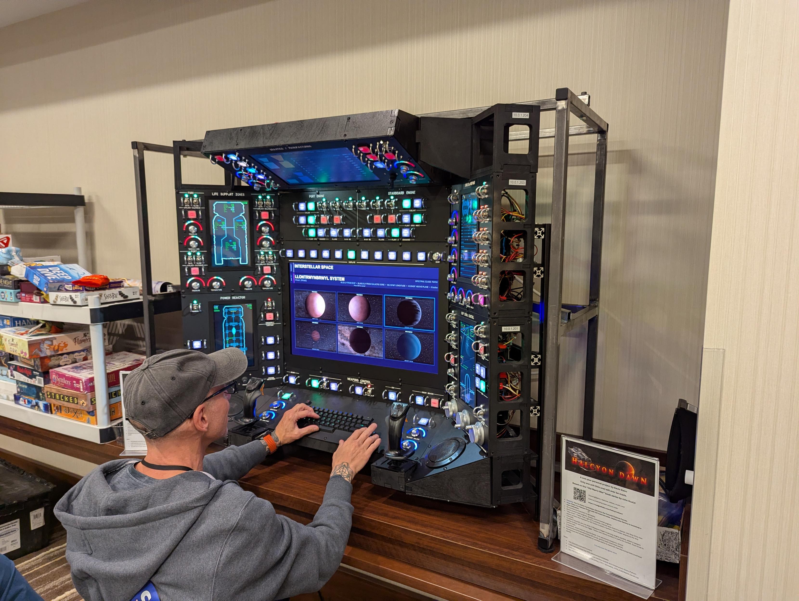 A man at a self made game console, 3 feet a side, buttons covered switches, joystick, and pedals.