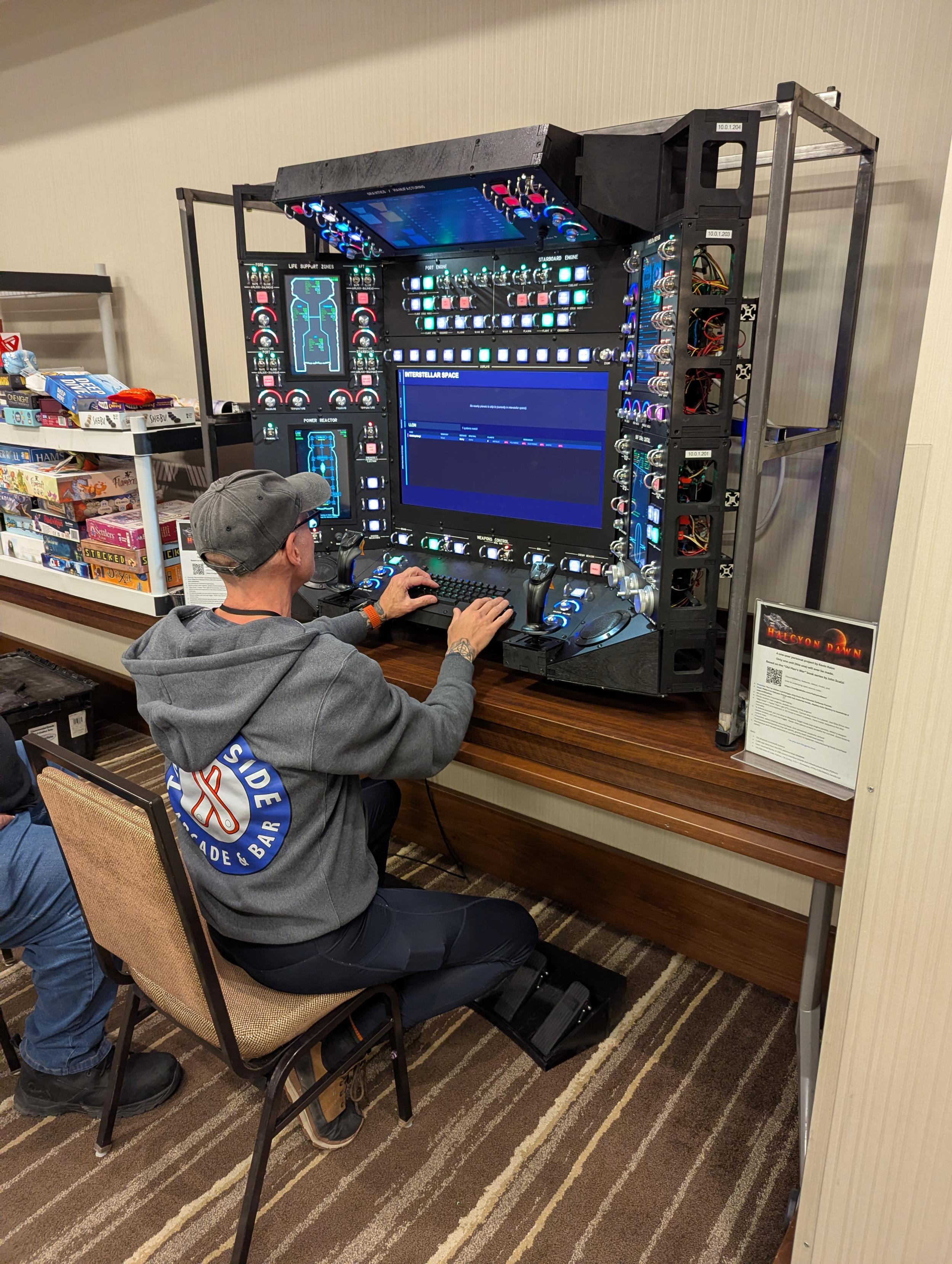 A man at a self made game console, 3 feet a side, buttons covered switches, joystick, and pedals.