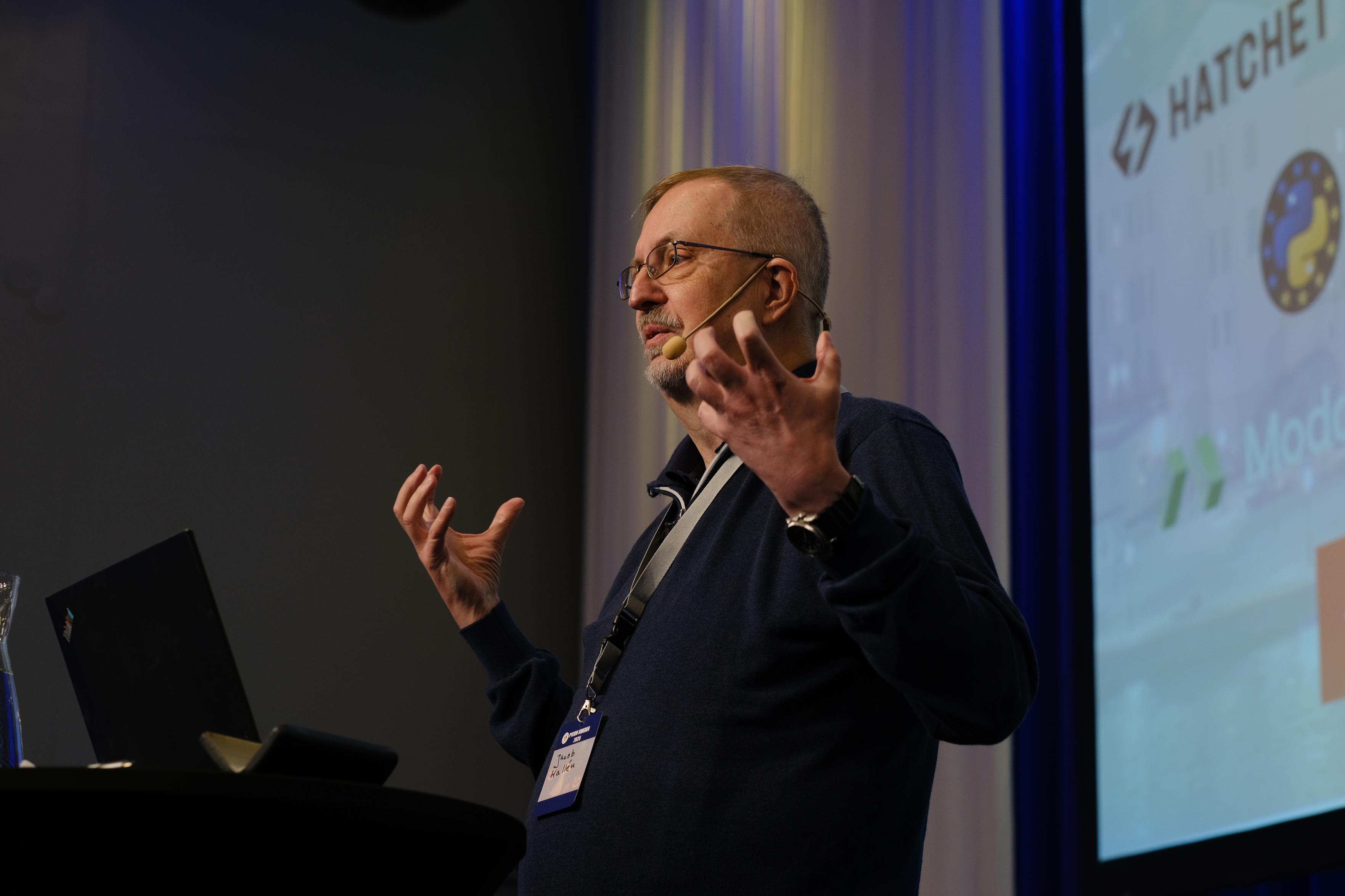 The image shows a man giving a presentation on a stage, likely at a technology conference.
**The Speaker:**
* He is a middle-aged man with short hair, a beard, and glasses.
* He is wearing a dark blue long-sleeved sweater or shirt.
* He has a headset microphone positioned near his mouth.
* He is wearing a lanyard around his neck with a conference badge attached.
* He is gesturing with both hands, palms facing somewhat inward, as if he is explaining a concept or emphasizing a point. He