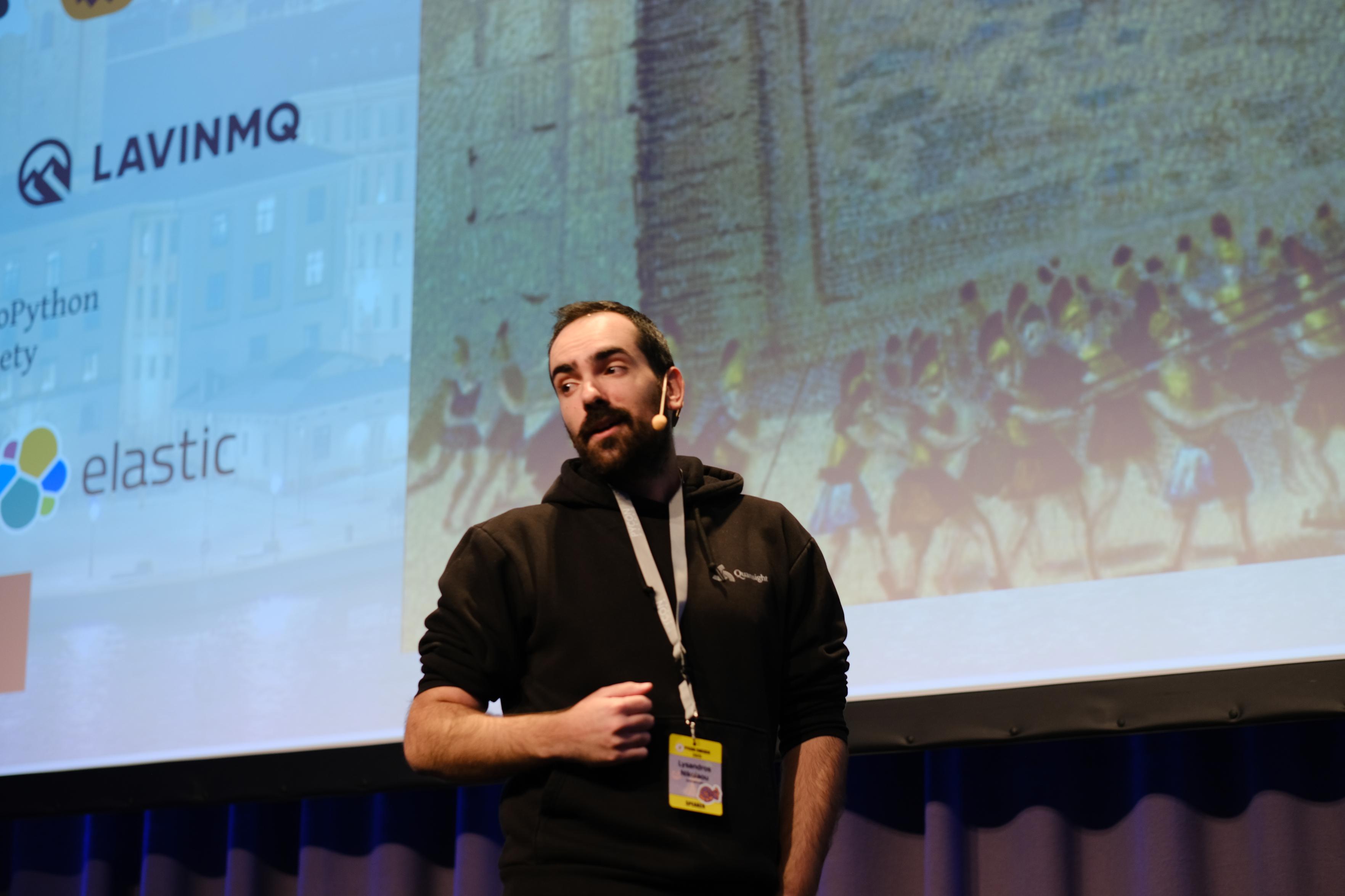 This image shows a man standing on a stage, likely giving a presentation or speech. He has short dark hair and a beard, and is wearing a black hooded sweatshirt. Around his neck is a lanyard with a yellow conference badge attached to it. He is also wearing a headset microphone.
Behind him is a large projection screen displaying a slide. On the left side of the screen, there are logos visible, including "LAVINMQ" and the logo for "elastic." The right side of the screen features a large, somewhat