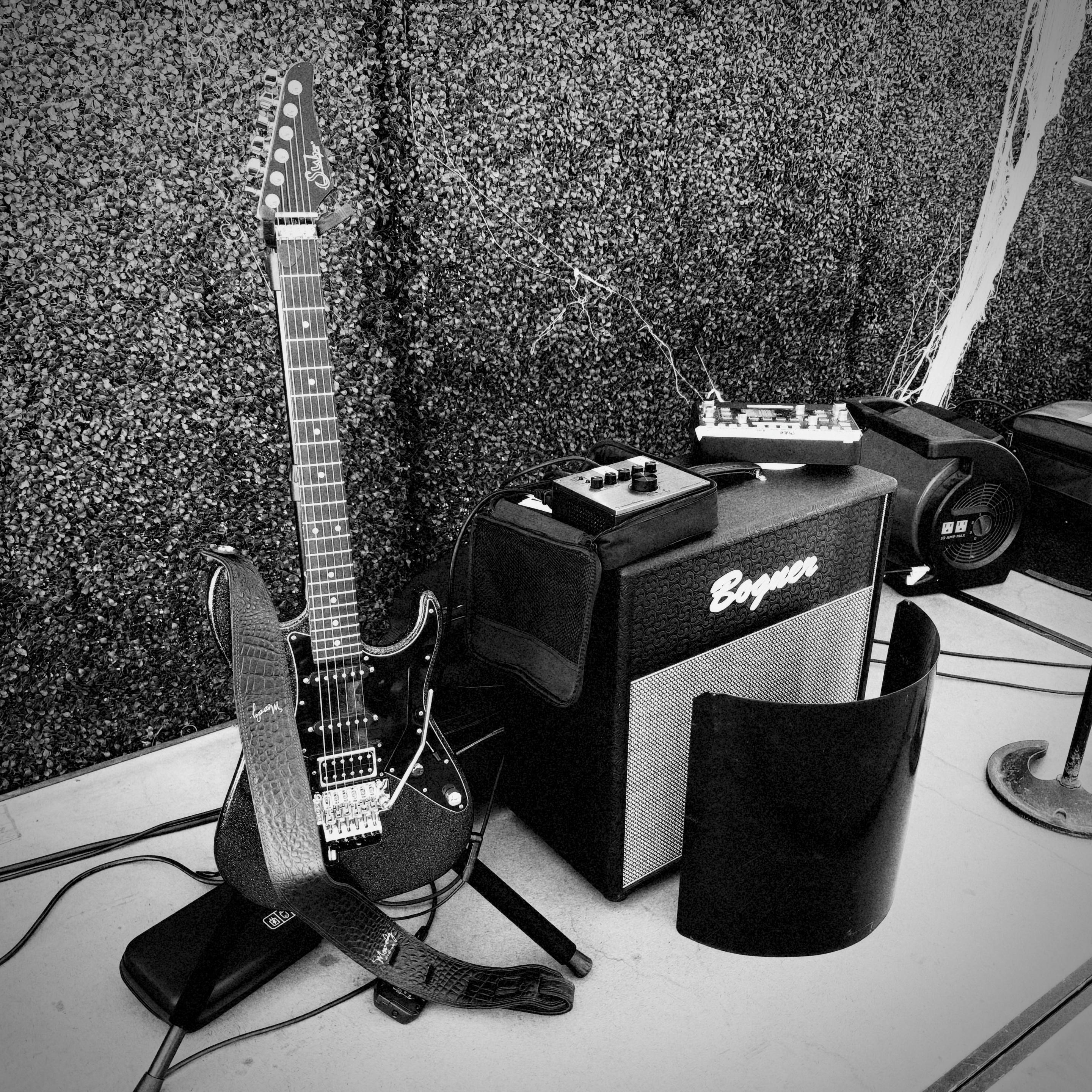 Black-and-white image of a black electric guitar on a stand beside a Bogner amplifier with pedals and cables on a floor.