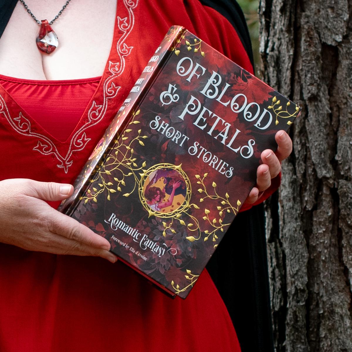 Person in a red embroidered dress holding a decorative hardcover book titled Of Blood & Petals: Short Stories — dark red cover with gold vine ornament and a small illustrated medallion, posed against a tree trunk.