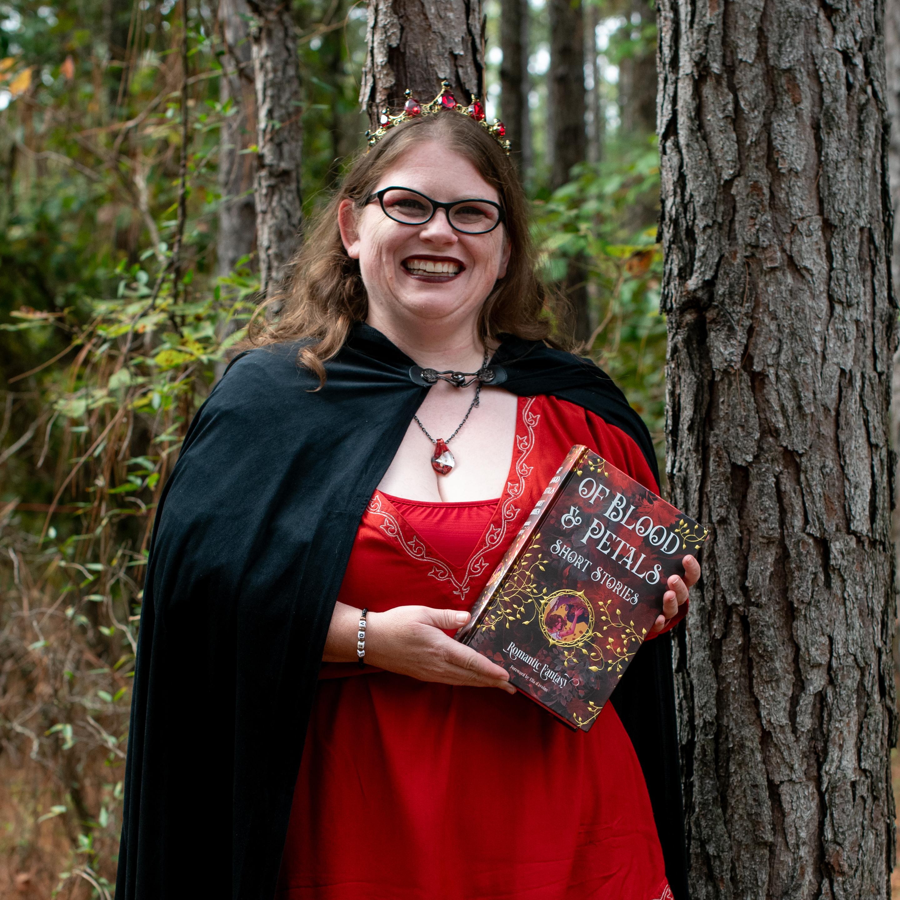 Smiling woman in a red dress and black cloak wearing a jeweled crown standing in a forest and holding a hardcover book titled "Of Blood & Petals," evoking a fantasy author or cosplay promotional portrait.