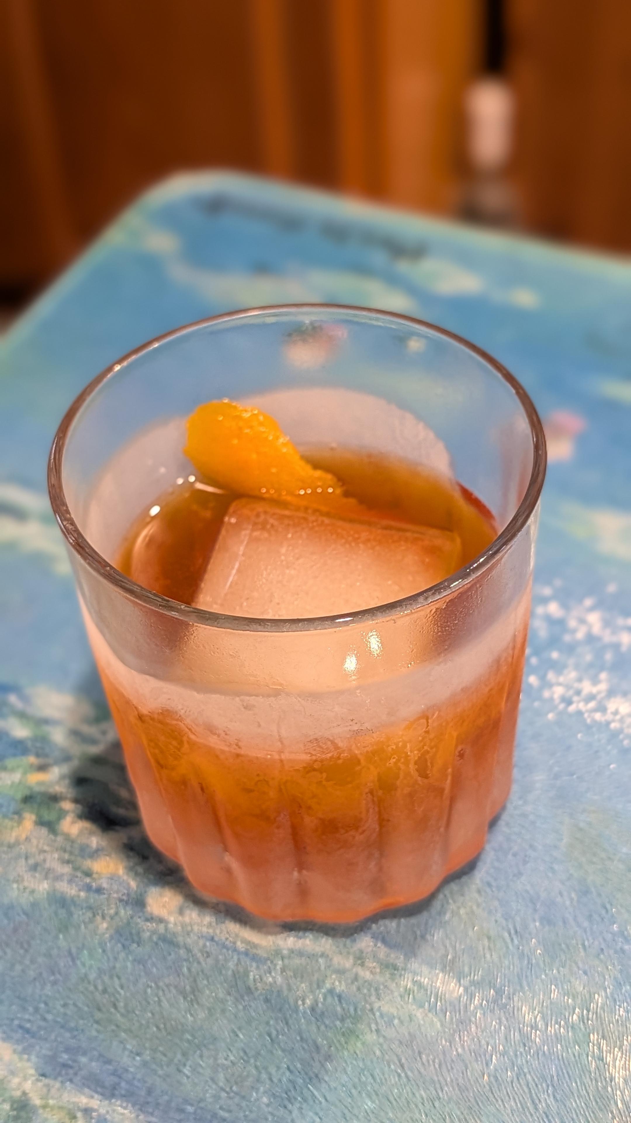 An orange cocktail featuring a large, clear square ice cube and an orange peel twist is served in a frosty ridged lowball glass. The glass rests on a textured blue and white patterned glass surface with a softly blurred background.